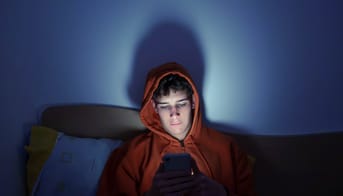 Teenage-dreng sidder alene i en sofa i mørket og kigger på sin telefon