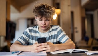 Teenagedreng sidder hjemme, kigger på telefon og laver lektier